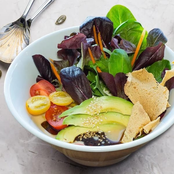 Colorful salad with mixed greens, cherry tomatoes, avocado, and sesame seeds in a white bowl.