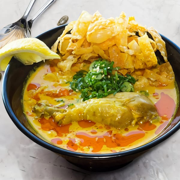 A vibrant bowl of Khao Soi with chicken, crispy noodles, and fresh scallions, garnished with lemon.