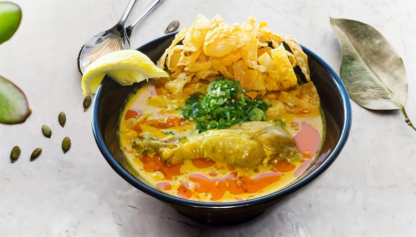 A vibrant bowl of Khao Soi with chicken, crispy noodles, and fresh scallions, garnished with lemon.